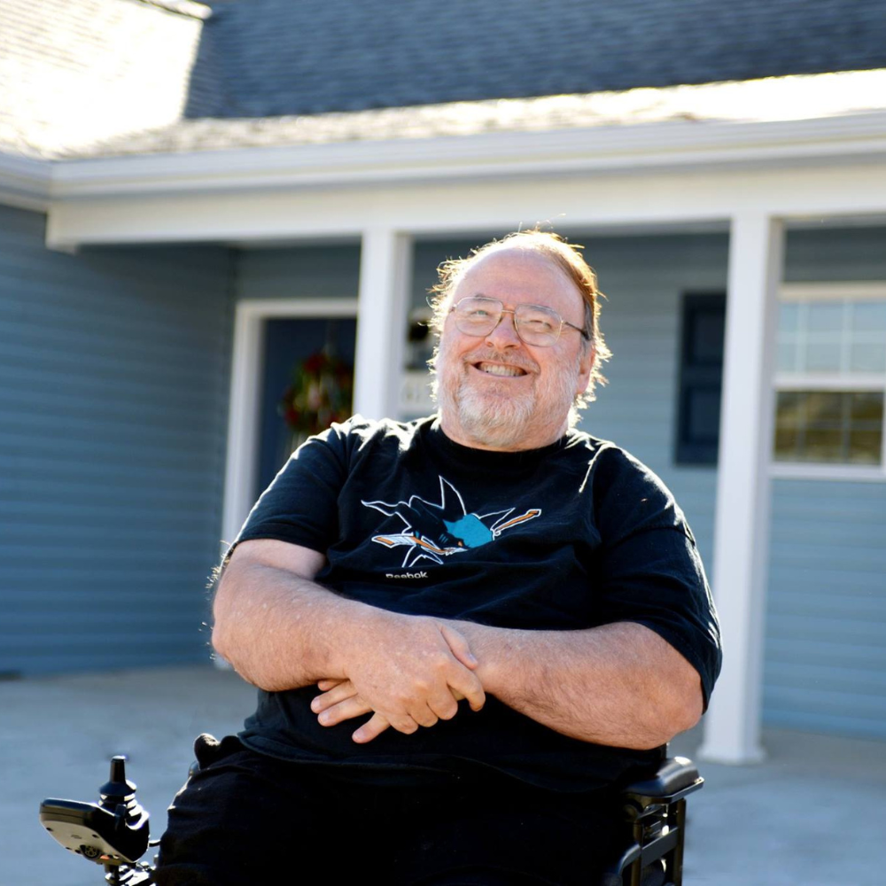 A middle-aged man is smiling and sitting in a wheelchair outside of a blue single-story.