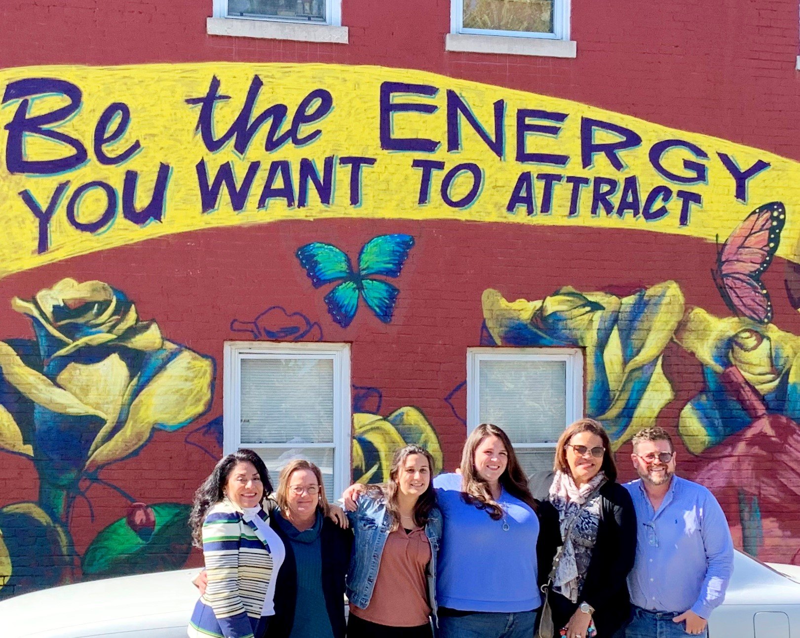 A group of six people are standing in front of a painted mural that says "Be the energy you want to attract". There are roses and butterflies on the mural.