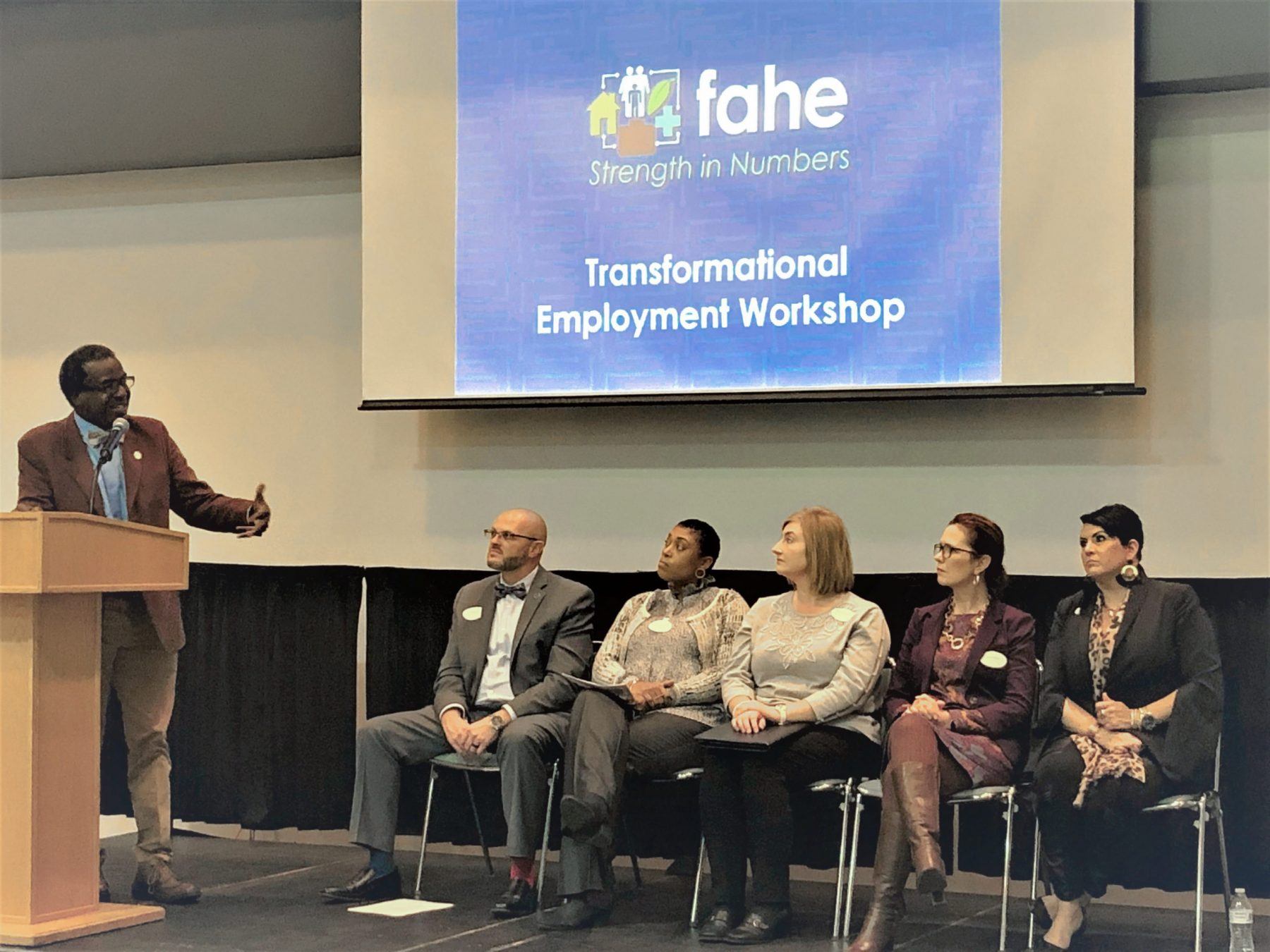 A group of five people are seated listening to a presenter speak at a podium. That is a Transformational Employment Workshop presentation screen behind them.