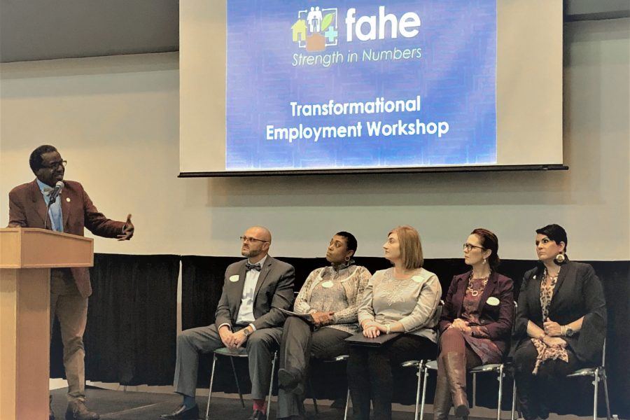 A group of five people are seated listening to a presenter speak at a podium. That is a Transformational Employment Workshop presentation screen behind them.