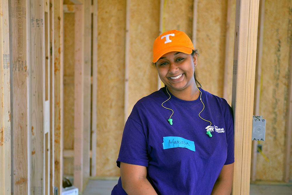 A woman is smiling and standing in a doorframe of an unfinished home. She is wearing a Habitat for Humanity t-shirt.