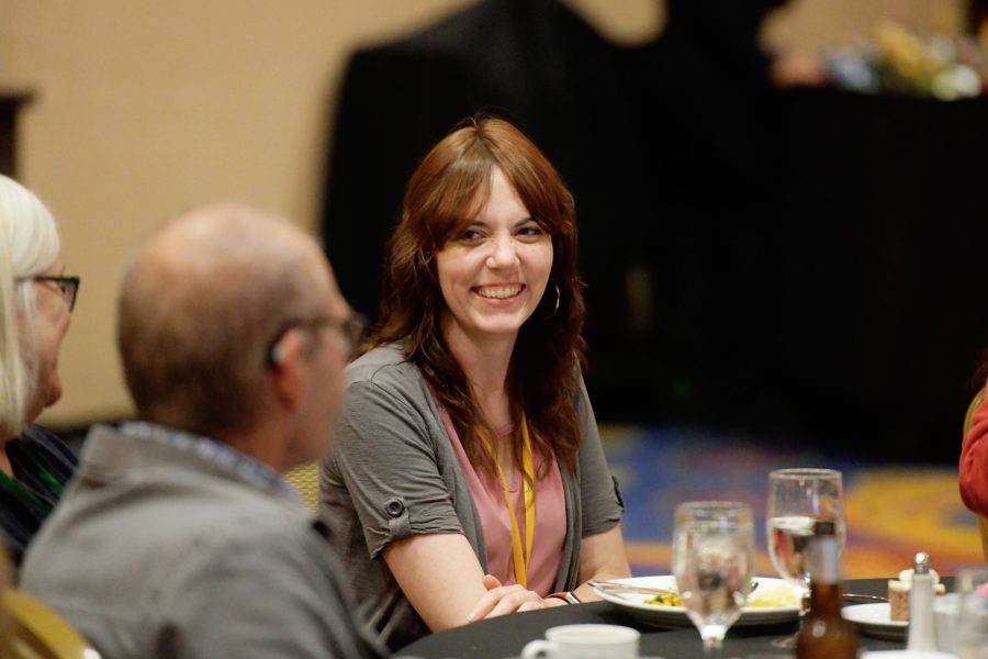 A woman is seated at a table smiling looking at someone else seated at the table. They are inside a ballroom at an annual event.