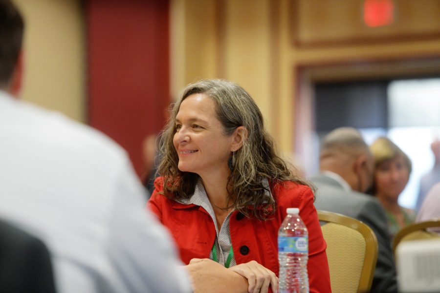 A woman is sitting at a table in a ballroom smiling. There are chairs and people sitting behind her.