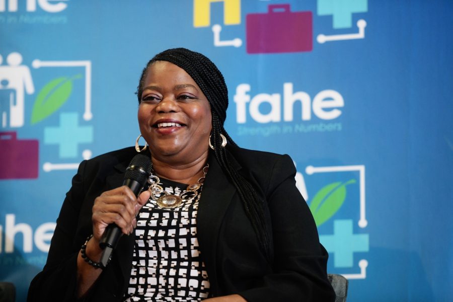 A woman of color is smiling and holding a microphone at an annual event. There is a Fahe step-and-repeat backdrop behind her.