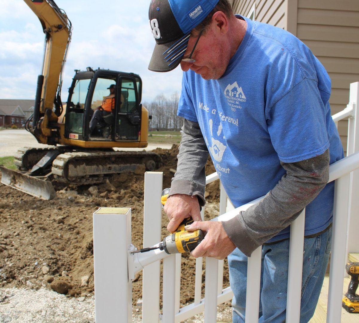 A man is drilling into a porch staircase railing outside. A small construction crane and dirt and rubble are behind him.