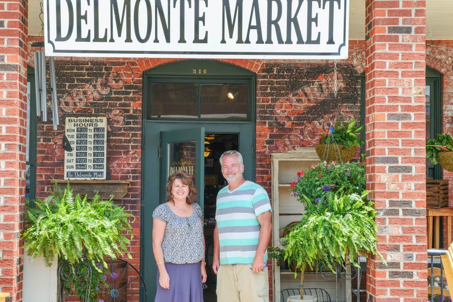 Two people are standing outside of the Delmonte Market. A green door and brick walls are behind them and there is an array of plants beside them.