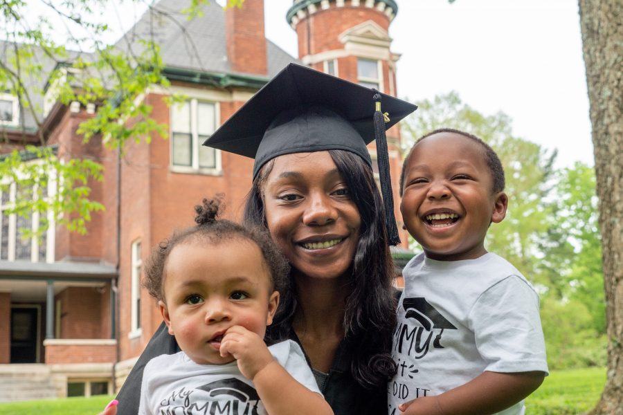 A mother is smiling holding her two smiling children outside. She is wearing a black university cap and gown.