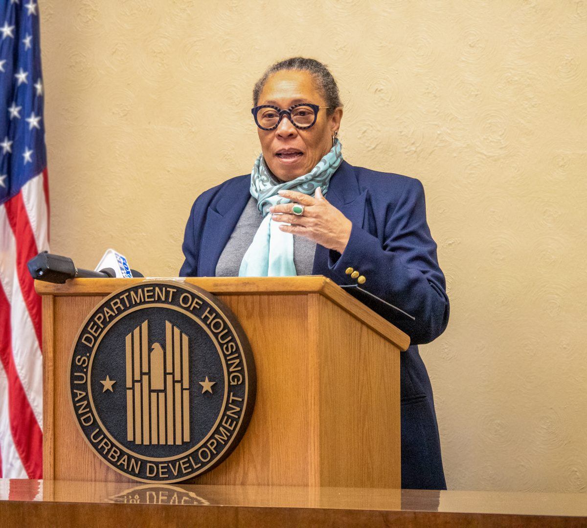 A woman is speaking at a podium that has U.S Department of Housing and Urban Development written on the front it. There is a tan wall behind her and an American flag.